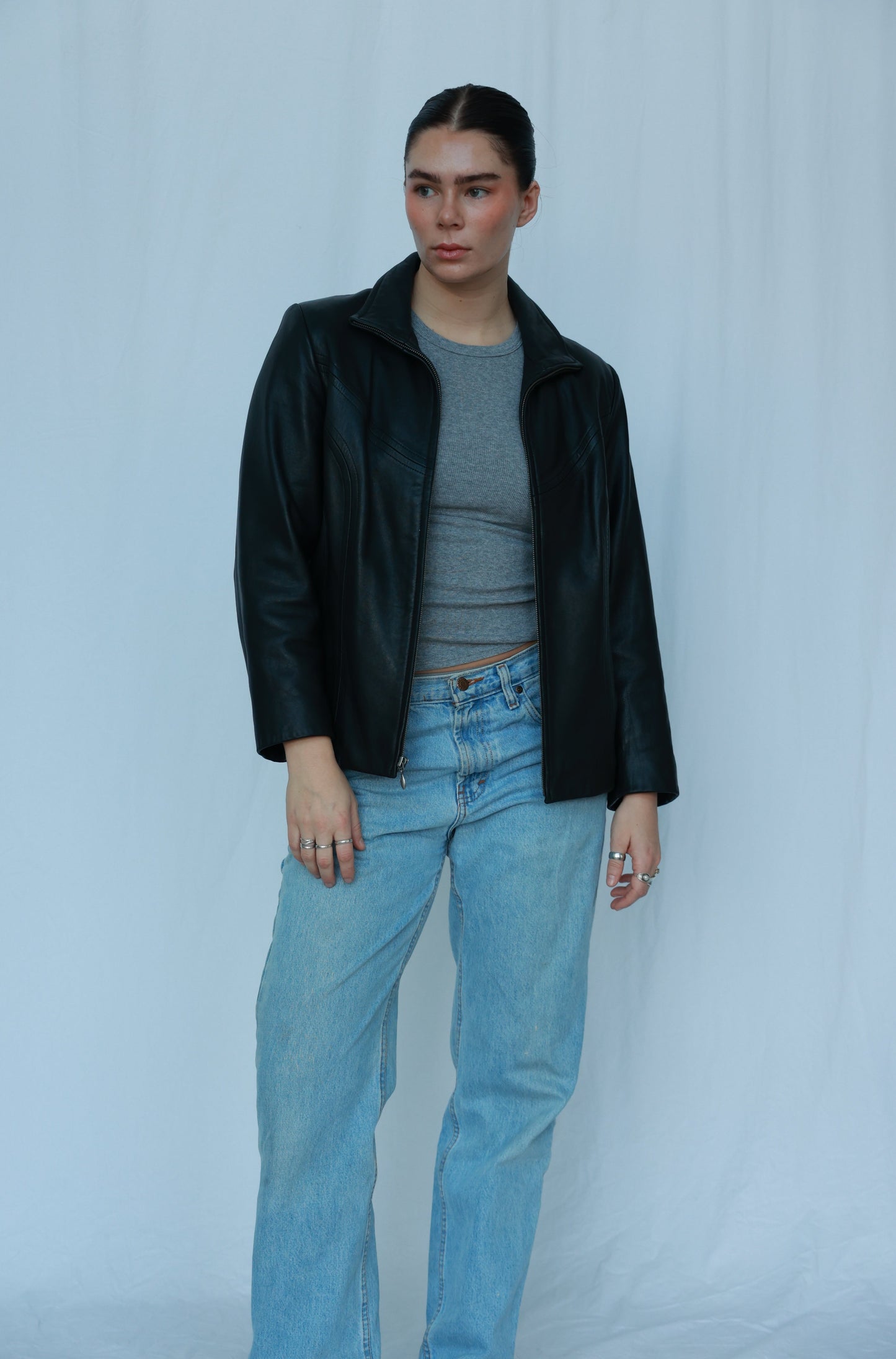 The image features a girl standing against a white backdrop. She's wearing a stylish black leather jacket over a grey tank top, paired with classic blue jeans. The ensemble exudes a trendy and casual vibe, creating a fashionable yet comfortable look.