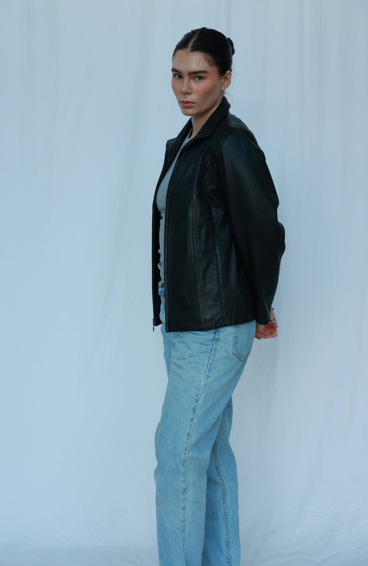 The image features a girl standing against a white backdrop. She's wearing a stylish black leather jacket over a grey tank top, paired with classic blue jeans. The ensemble exudes a trendy and casual vibe, creating a fashionable yet comfortable look.