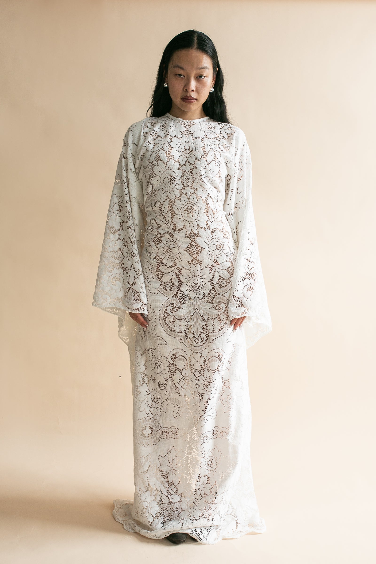 A model wearing a long-sleeved, high-neck white lace dress with intricate floral embroidery, standing against a solid beige studio background.
