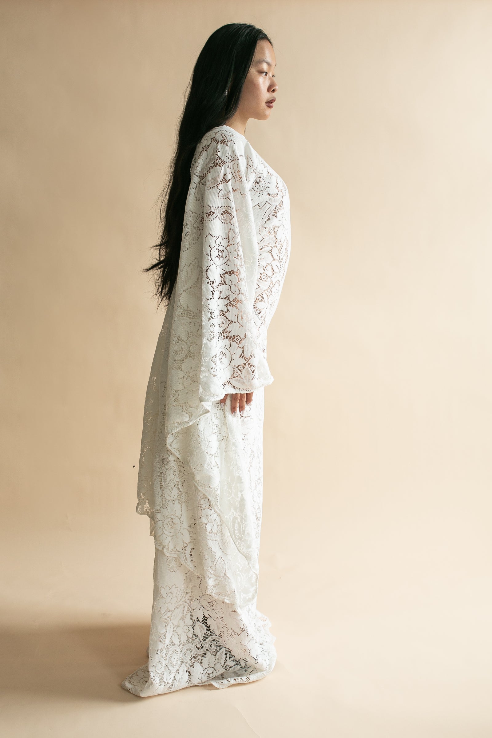 A woman in profile wearing a long-sleeved, floor-length white lace dress against a solid beige studio background.