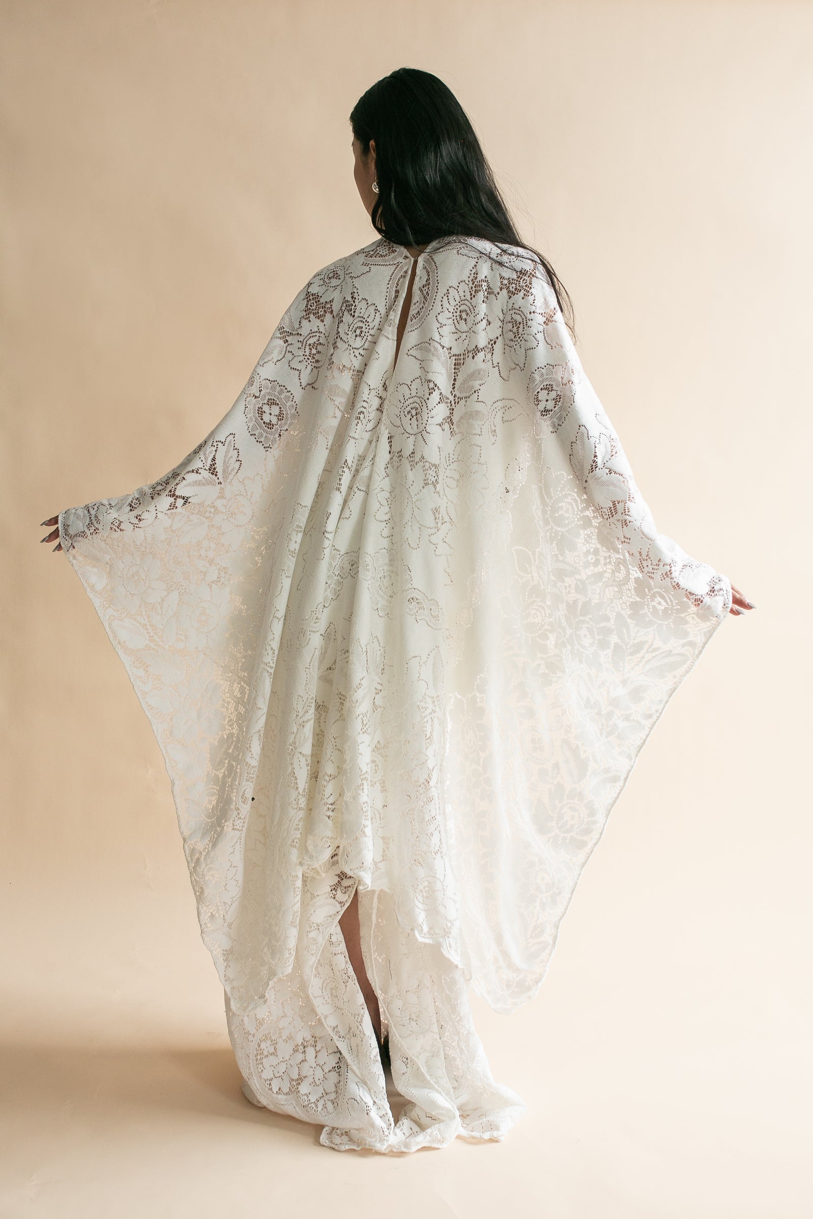 A woman viewed from behind wearing a flowing ivory lace robe or caftan with wide draped sleeves, posed against a plain warm beige studio background.