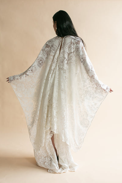 A woman viewed from behind wearing a flowing ivory lace robe or caftan with wide draped sleeves, posed against a plain warm beige studio background.